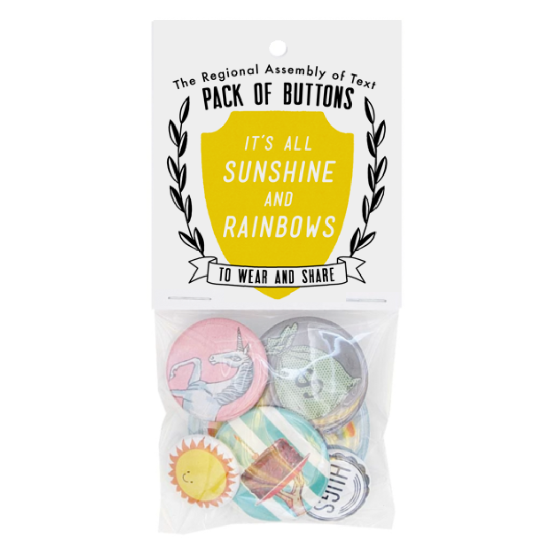 A clear plastic package containing six colorful pin-back buttons. The header card of the package reads "The Regional Assembly of Text PACK OF BUTTONS" and features a yellow shield with "IT'S ALL SUNSHINE AND RAINBOWS" inside, and "TO WEAR AND SHARE" below. Visible buttons include a unicorn, a sun, and a design with stripes and a building.