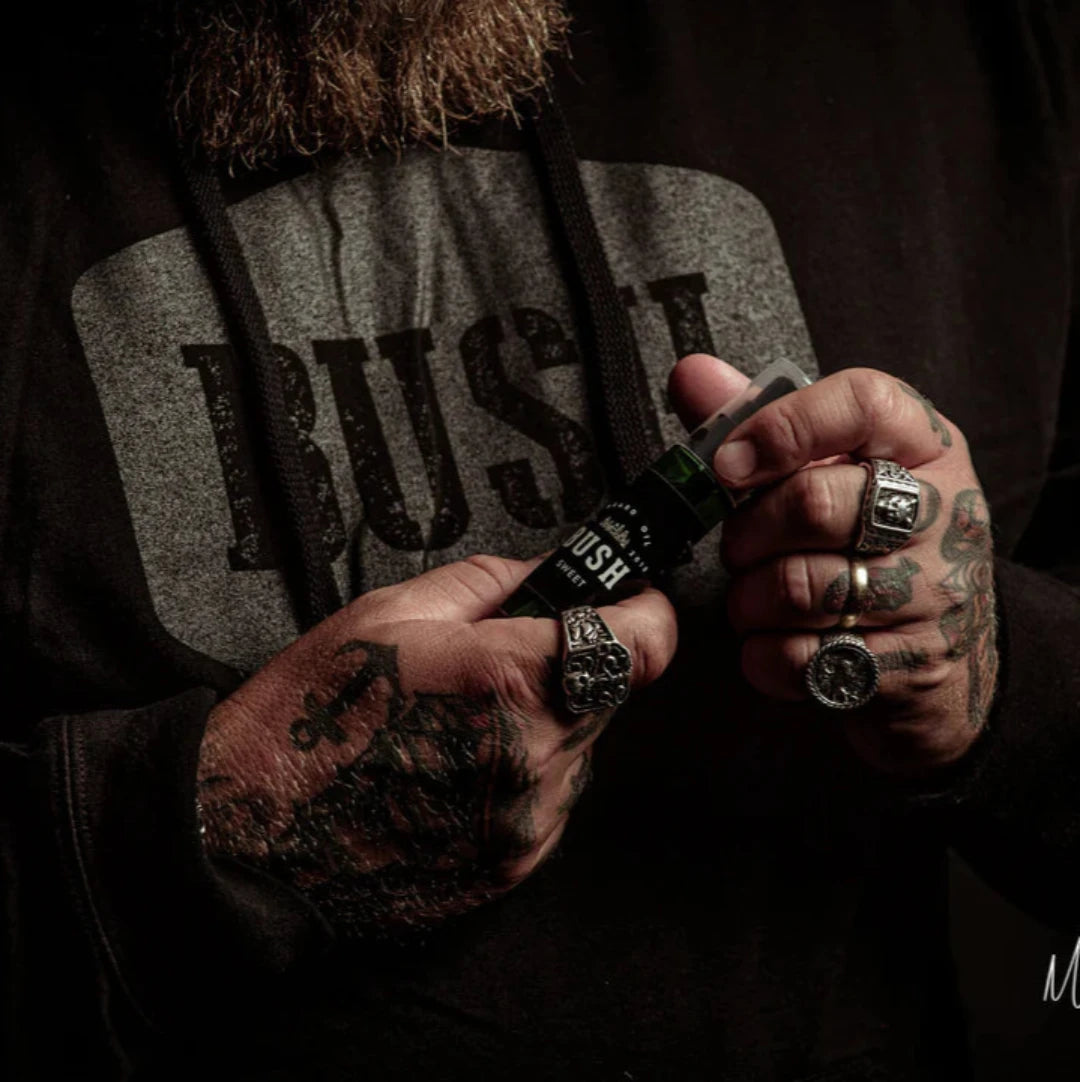 A dark green glass bottle with a black pump dispenser. The bottle is labeled "BUSH" and "SWEET BEARD OIL." The background is black.