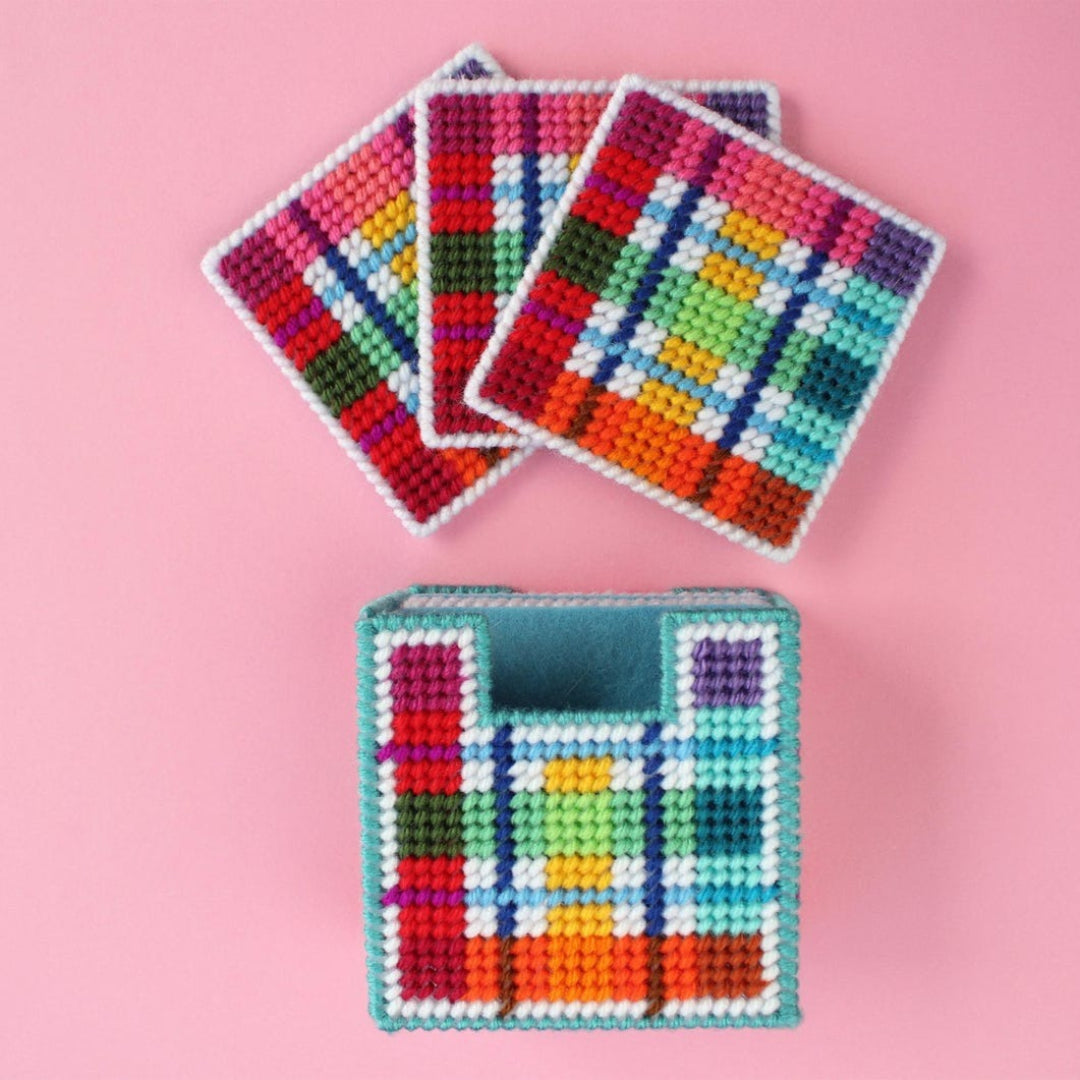 An overhead shot of a needlepoint coaster kit on a pink background. Four finished coasters with a colourful plaid pattern are fanned out at the top, while a matching square coaster holder is at the bottom.