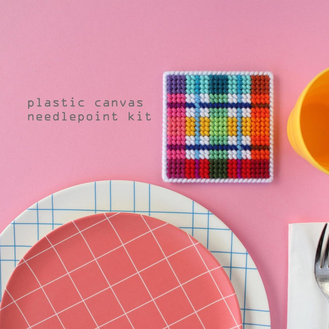 An overhead shot of a needlepoint coaster kit on a pink background. Four finished coasters with a colourful plaid pattern are fanned out at the top, while a matching square coaster holder is at the bottom.