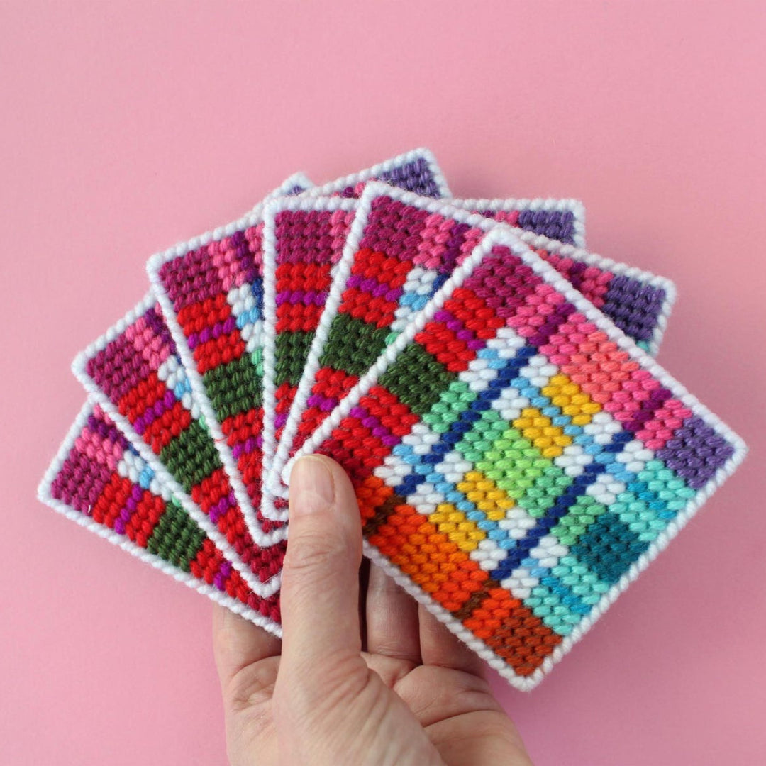 An overhead shot of a needlepoint coaster kit on a pink background. Four finished coasters with a colourful plaid pattern are fanned out at the top, while a matching square coaster holder is at the bottom.