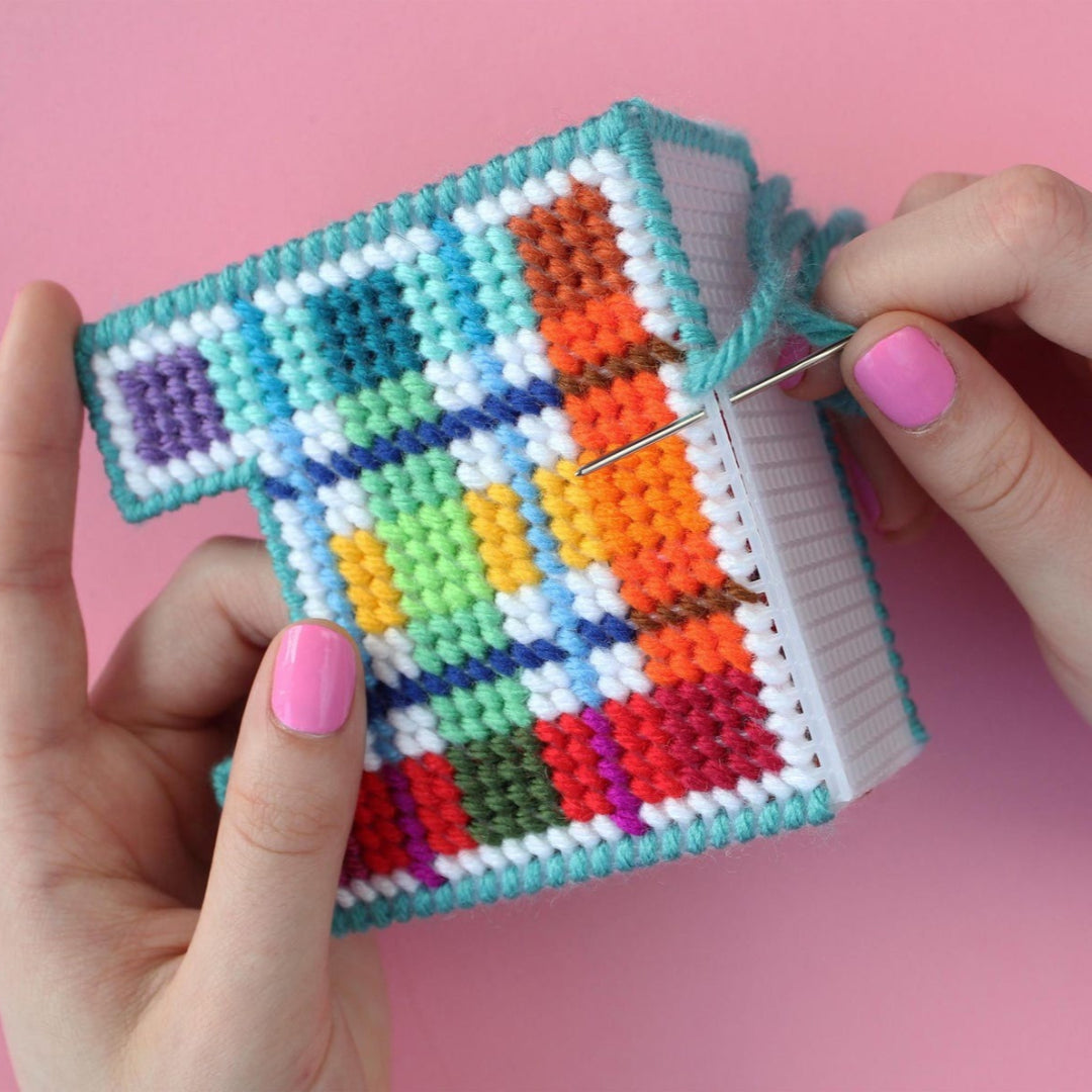 An overhead shot of a needlepoint coaster kit on a pink background. Four finished coasters with a colourful plaid pattern are fanned out at the top, while a matching square coaster holder is at the bottom.