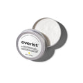 A small, open, silver metal tin of Everist's "The Fragrance-Free Shampoo Concentrate Mini," revealing a white, concentrated product inside. The tin's lid is propped against the base, with text indicating it is dermatologist-developed and contains 20ml of product.