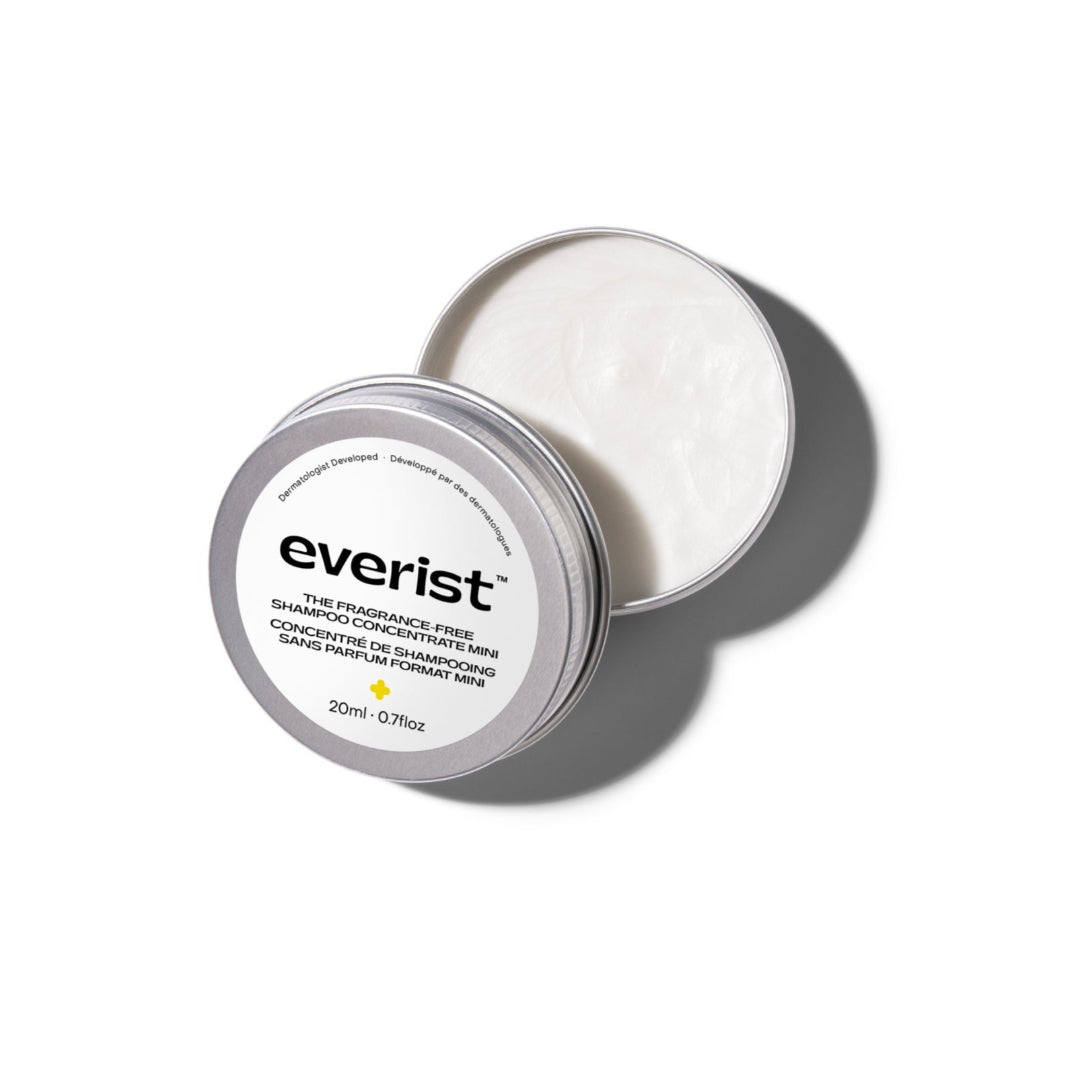 A small, open, silver metal tin of Everist's "The Fragrance-Free Shampoo Concentrate Mini," revealing a white, concentrated product inside. The tin's lid is propped against the base, with text indicating it is dermatologist-developed and contains 20ml of product.
