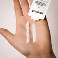 Hand holding a small amount of cream with a visible brand label on a neutral background