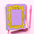 A notepad designed to look like the purple door from the TV show Friends, complete with the iconic yellow picture frame. The text at the top reads "THE ONE WHERE I GET STUFF DONE." A pink pen is next to the notepad on a pink background.