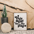 Decorative sign with 'This Must be the Place' text, potted plant, and round pillow against a wooden background.