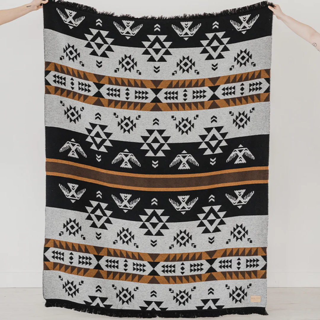 The Thunderbird Reversible Eco-Friendly Blanket by Mini Tipi, featuring bold Indigenous-inspired Thunderbird and geometric patterns in black, grey, and brown, held up by two hands. This sustainable and culturally significant blanket is available in Ottawa, Ontario, Canada.