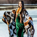 The Thunderbird Reversible Eco-Friendly Blanket by Mini Tipi, featuring bold Indigenous-inspired Thunderbird and geometric patterns in black, grey, and brown, hanging on a shoulders and arms of a person. This sustainable and culturally significant blanket is available in Ottawa, Ontario, Canada.