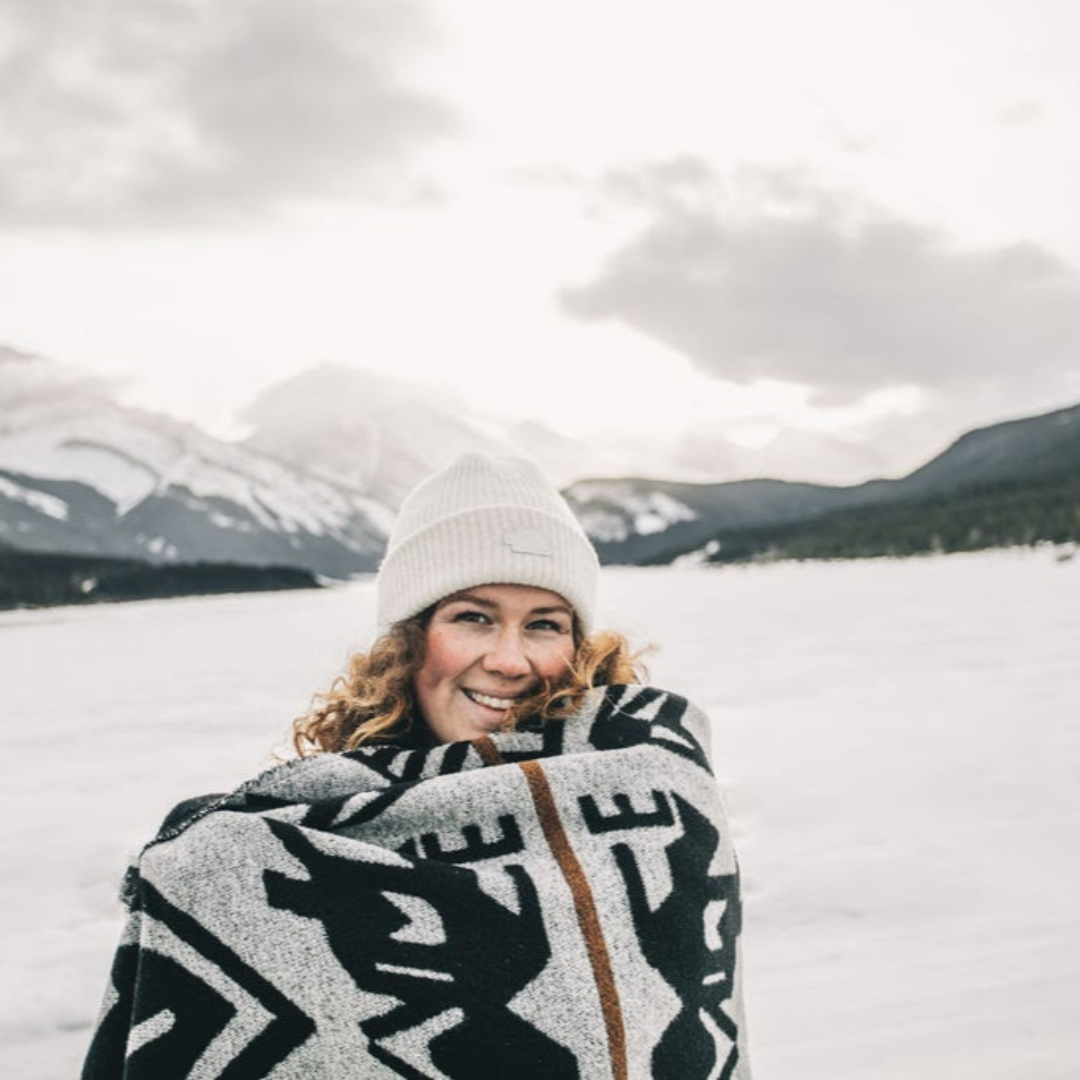 The Thunderbird Reversible Eco-Friendly Blanket by Mini Tipi, featuring bold Indigenous-inspired Thunderbird and geometric patterns in black, grey, and brown, wrapped around a person in the mountains. This sustainable and culturally significant blanket is available in Ottawa, Ontario, Canada.
