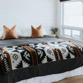 The Thunderbird Reversible Eco-Friendly Blanket by Mini Tipi, featuring bold Indigenous-inspired Thunderbird and geometric patterns in black, grey, and brown, laying on a bed. This sustainable and culturally significant blanket is available in Ottawa, Ontario, Canada.