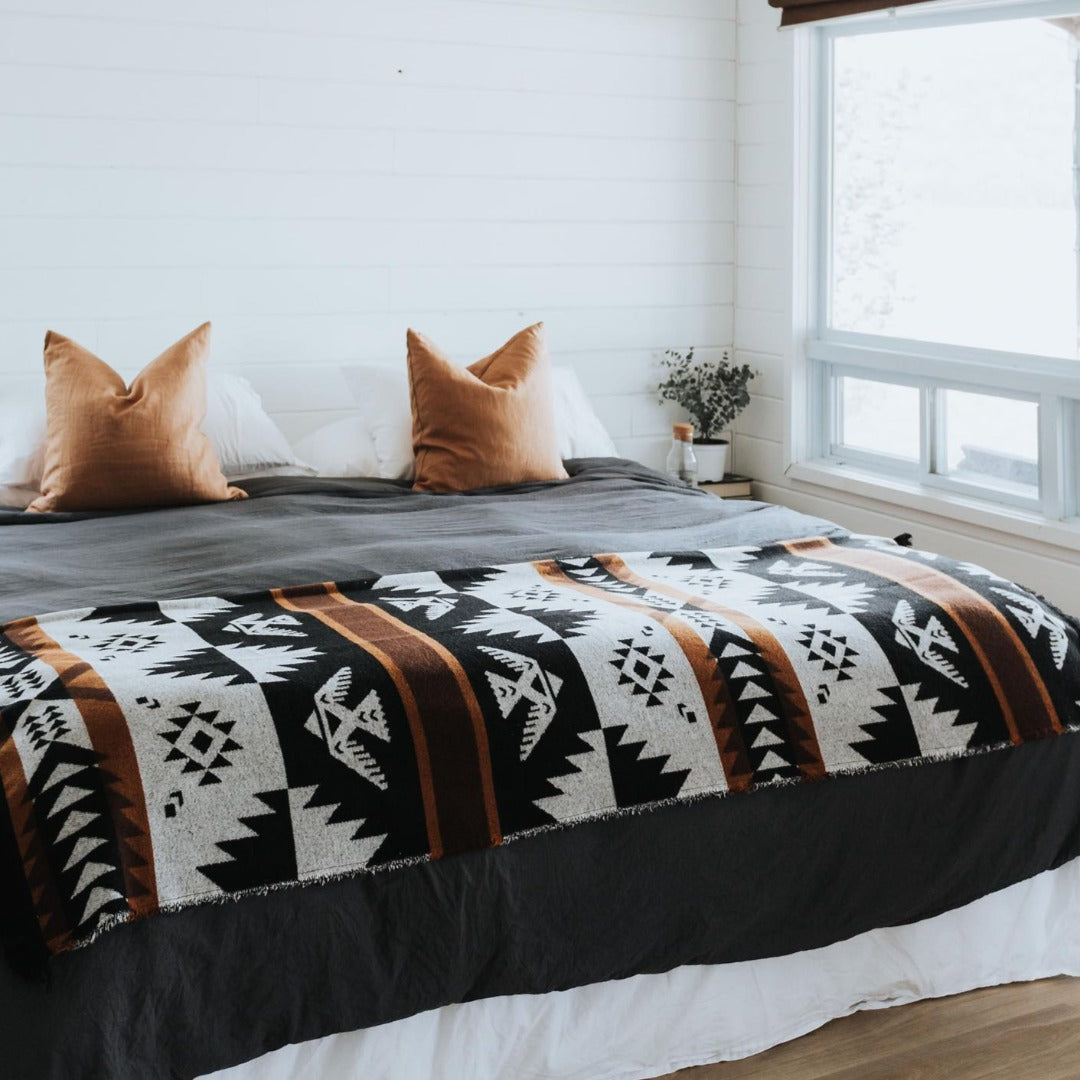 The Thunderbird Reversible Eco-Friendly Blanket by Mini Tipi, featuring bold Indigenous-inspired Thunderbird and geometric patterns in black, grey, and brown, laying on a bed. This sustainable and culturally significant blanket is available in Ottawa, Ontario, Canada.
