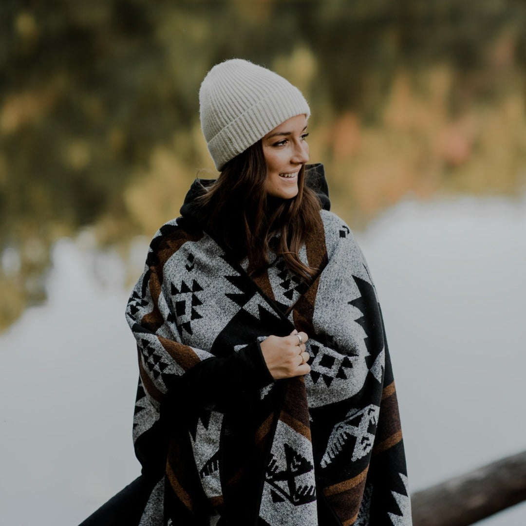 The Thunderbird Reversible Eco-Friendly Blanket by Mini Tipi, featuring bold Indigenous-inspired Thunderbird and geometric patterns in black, grey, and brown, wrapped around a person. This sustainable and culturally significant blanket is available in Ottawa, Ontario, Canada.