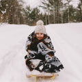 The Thunderbird Reversible Eco-Friendly Blanket by Mini Tipi, featuring bold Indigenous-inspired Thunderbird and geometric patterns in black, grey, and brown, wrapped around a young child riding in a small sled. This sustainable and culturally significant blanket is available in Ottawa, Ontario, Canada.