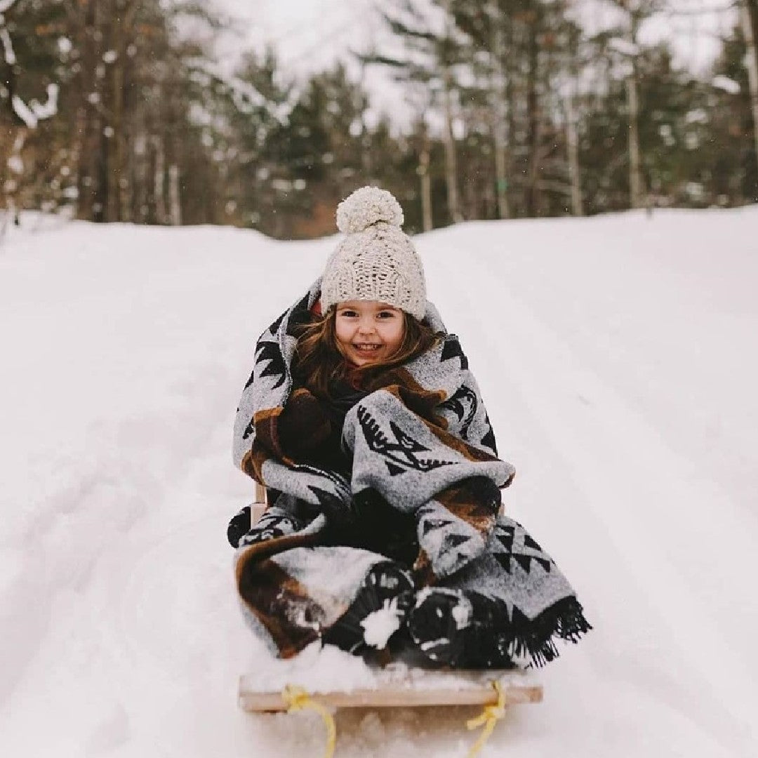 The Thunderbird Reversible Eco-Friendly Blanket by Mini Tipi, featuring bold Indigenous-inspired Thunderbird and geometric patterns in black, grey, and brown, wrapped around a young child riding in a small sled. This sustainable and culturally significant blanket is available in Ottawa, Ontario, Canada.