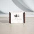A bar of Aide Bodycare Toasted Marshmallow Soap, featuring a dark brown color and a paper label with the 'aide' logo and 'Toasted Marshmallow' text, displayed on a light-colored surface with a shadow overlay. The soap is natural and handmade, available in Ottawa, Ontario, Canada.