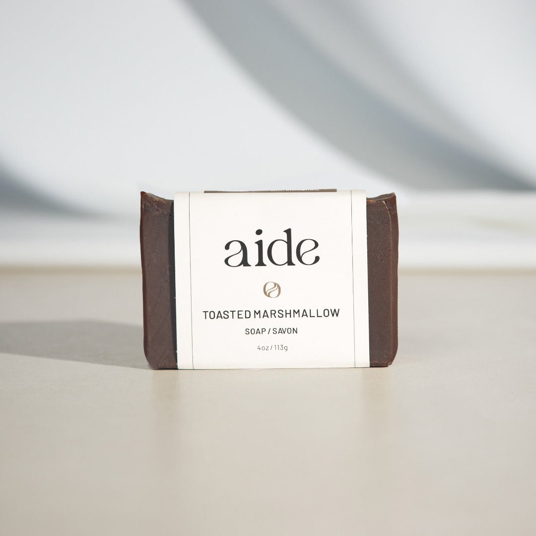 A bar of Aide Bodycare Toasted Marshmallow Soap, featuring a dark brown color and a paper label with the 'aide' logo and 'Toasted Marshmallow' text, displayed on a light-colored surface with a shadow overlay. The soap is natural and handmade, available in Ottawa, Ontario, Canada.