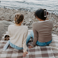 Two women sitting on a checkered blanket by the water.