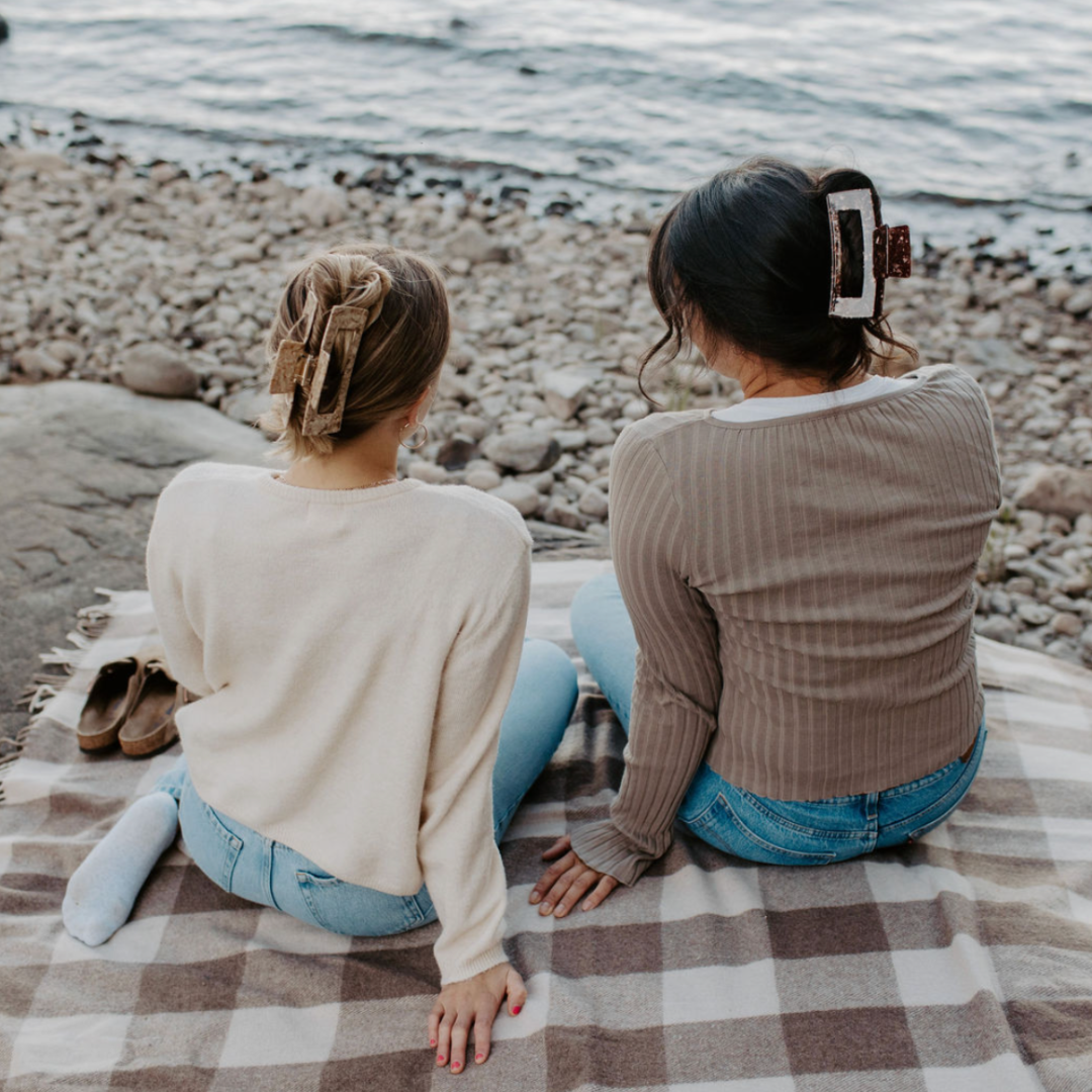 Two women sitting on a checkered blanket by the water.