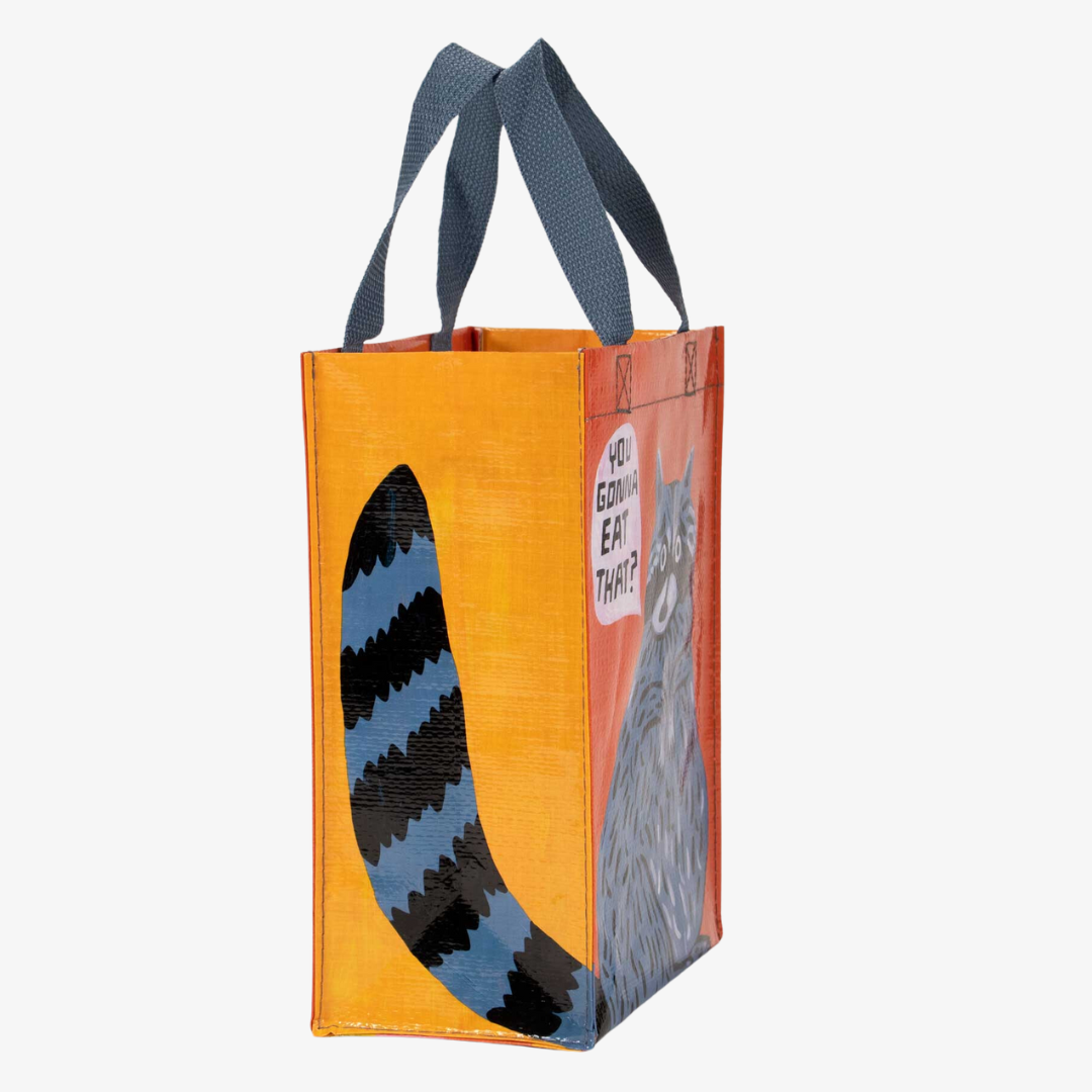A reusable tote bag with a playful design. The bag is orange and yellow with a blue raccoon in the center. The raccoon is holding a piece of food and has a speech bubble above its head that says "YOU GONNA EAT THAT?" The bag has blue handles.