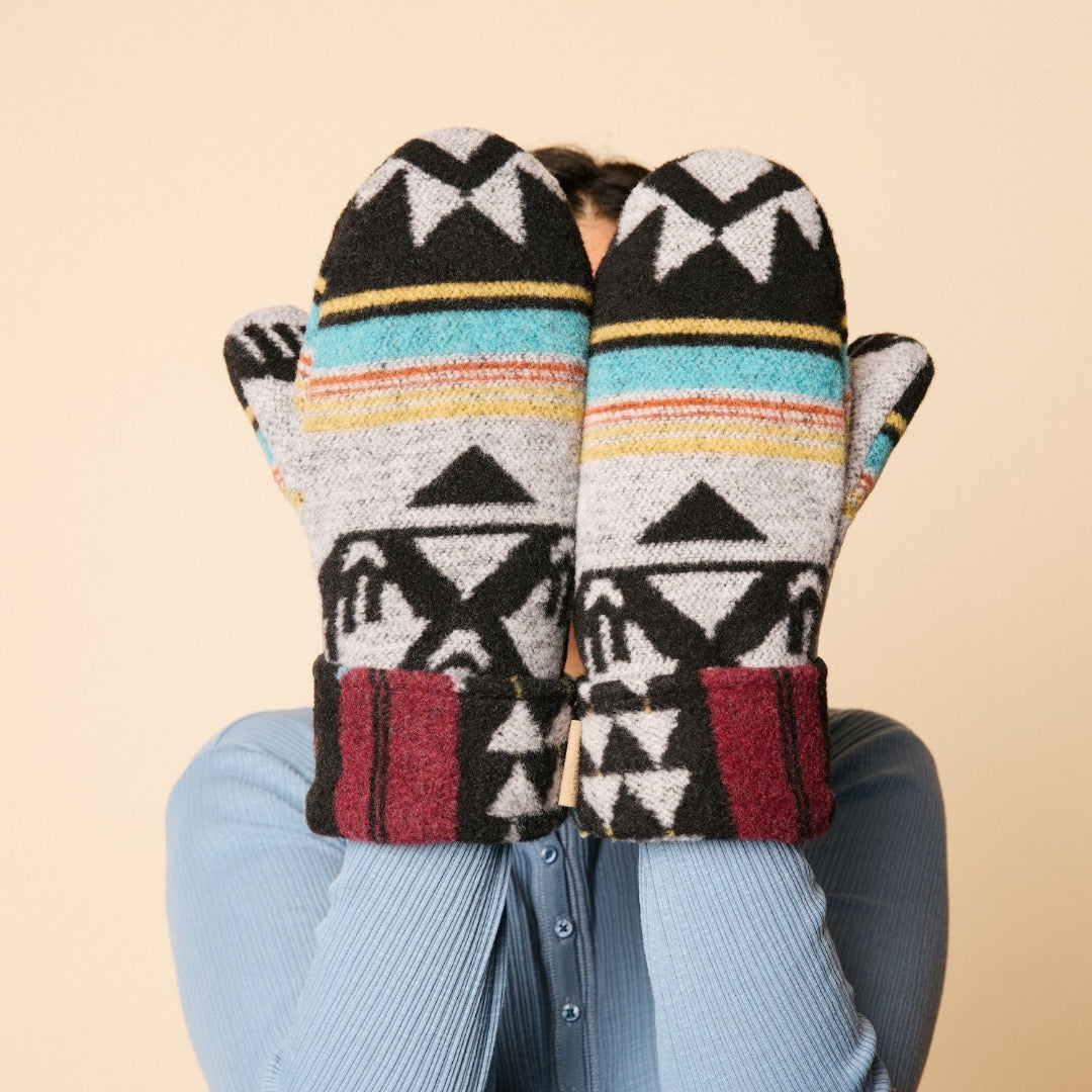 Pair of patterned mittens held by a person wearing a blue sweater against a beige background