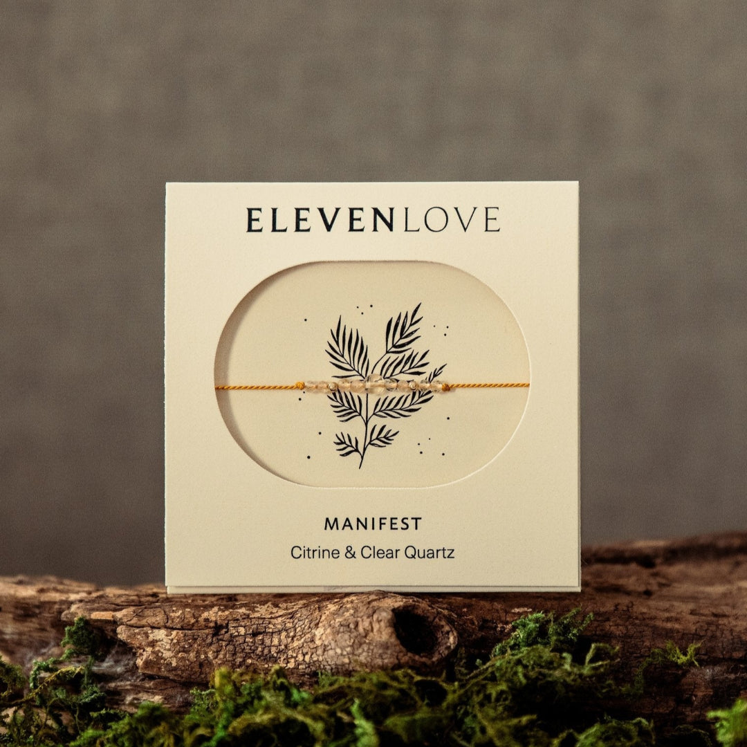 Eleven LoveManifest product packaging on a wooden surface with moss, against a brown background
