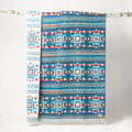 The Nibi Reversible Blanket by Mini Tipi, featuring bold Indigenous-inspired geometric patterns in teal, red, brown, and white, hanging from a natural branch. This reversible and stylish blanket is available in Ottawa, Ontario, Canada.