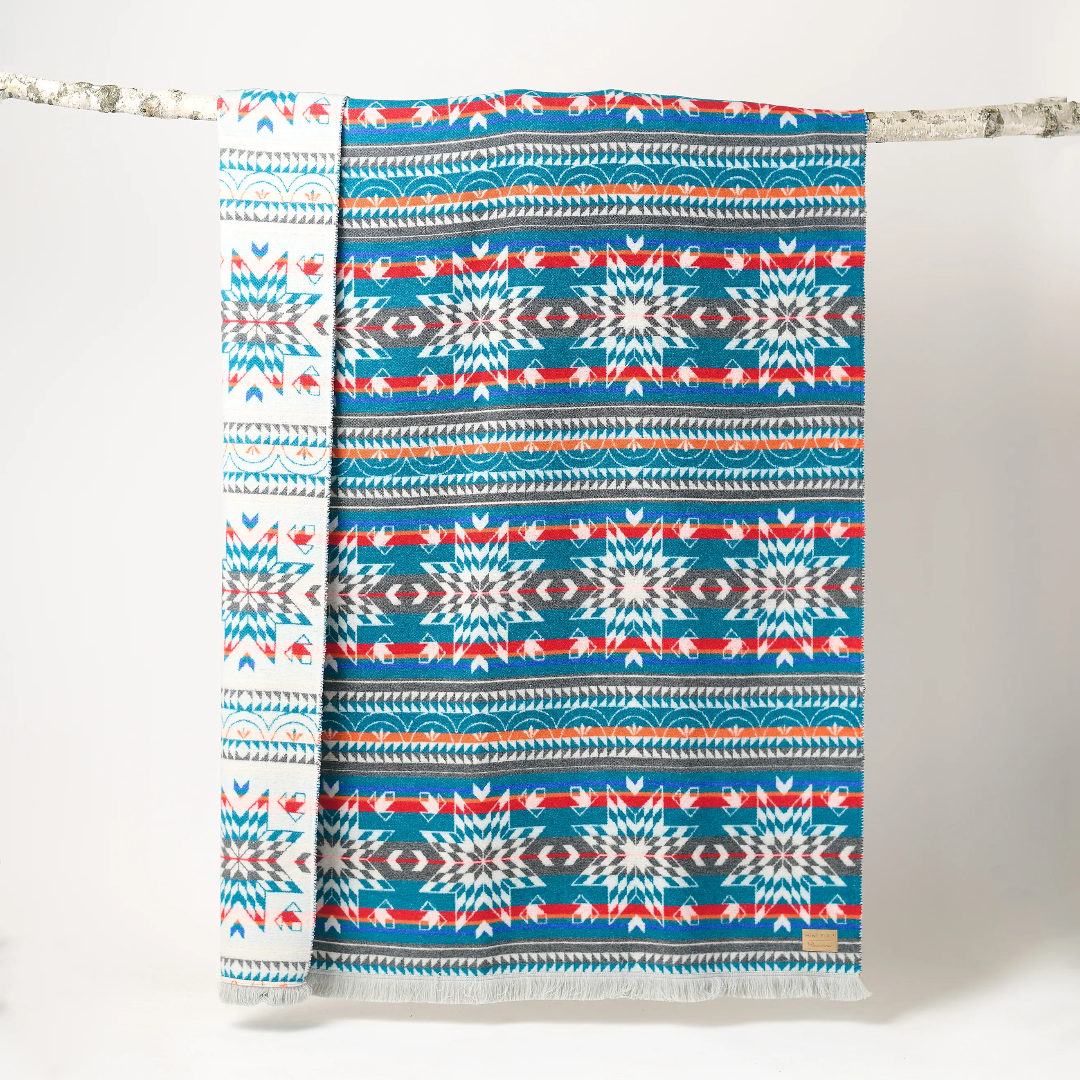 The Nibi Reversible Blanket by Mini Tipi, featuring bold Indigenous-inspired geometric patterns in teal, red, brown, and white, hanging from a natural branch. This reversible and stylish blanket is available in Ottawa, Ontario, Canada.