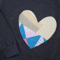 a grey sweatshirt with a large, heart-shaped patch. The patch is made from various fabrics with various patterns, sewn together in a patchwork style.