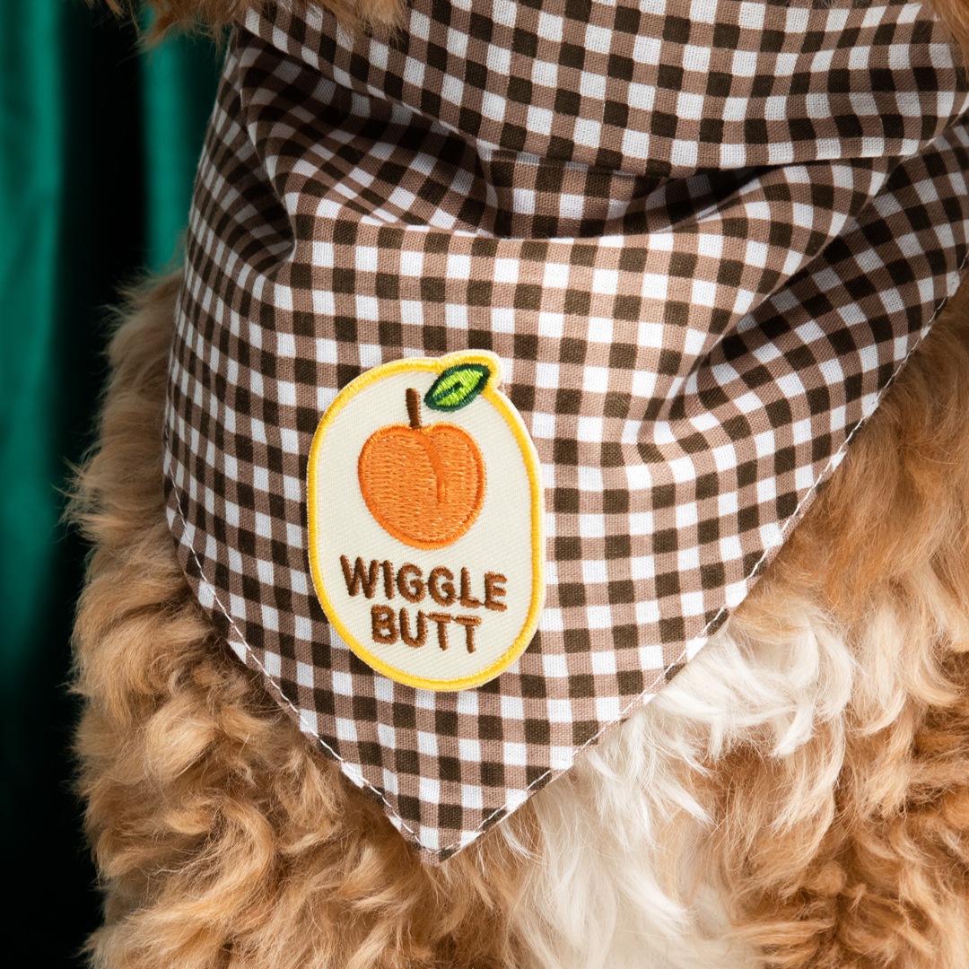 Dog wearing a checkered bandana with a patch labeled 'Wiggle Butt' on a brown background.