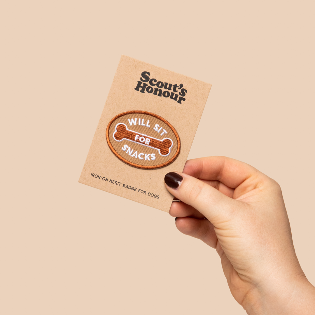 Hand holding a 'Scout's Honour' patch with a dog bone design on a beige background