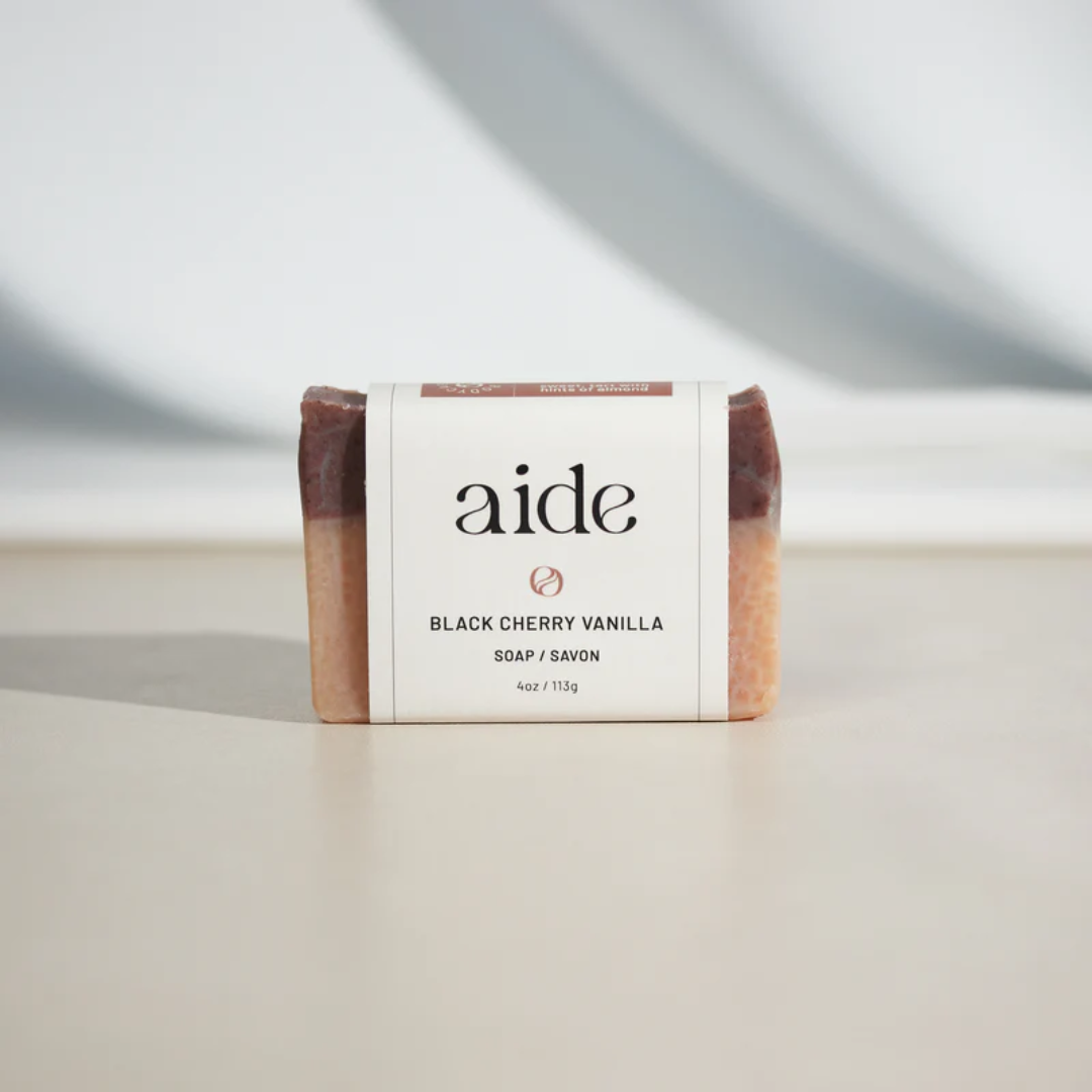 A bar of Aide Bodycare Black Cherry Vanilla Soap, featuring a layered light beige and dark red-brown color and a paper label with the 'aide' logo and 'Black Cherry Vanilla' text, displayed on a light-colored surface with a shadow overlay. The soap is natural and handmade, available in Ottawa, Ontario, Canada.