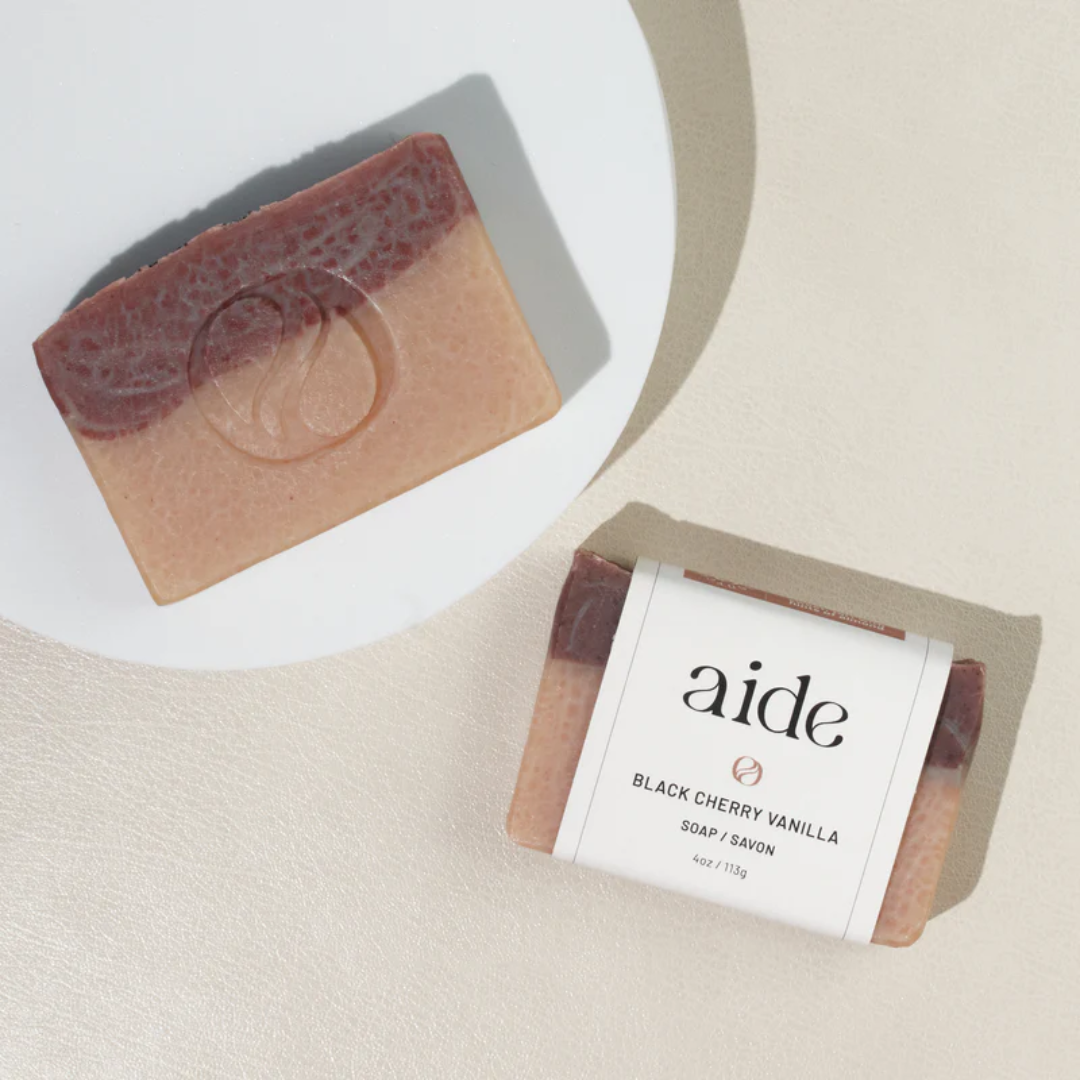 A bar of Aide Bodycare Black Cherry Vanilla Soap, featuring a layered light beige and dark red-brown color and a paper label with the 'aide' logo and 'Black Cherry Vanilla' text, displayed on a light-colored surface with a shadow overlay. The soap is natural and handmade, available in Ottawa, Ontario, Canada.