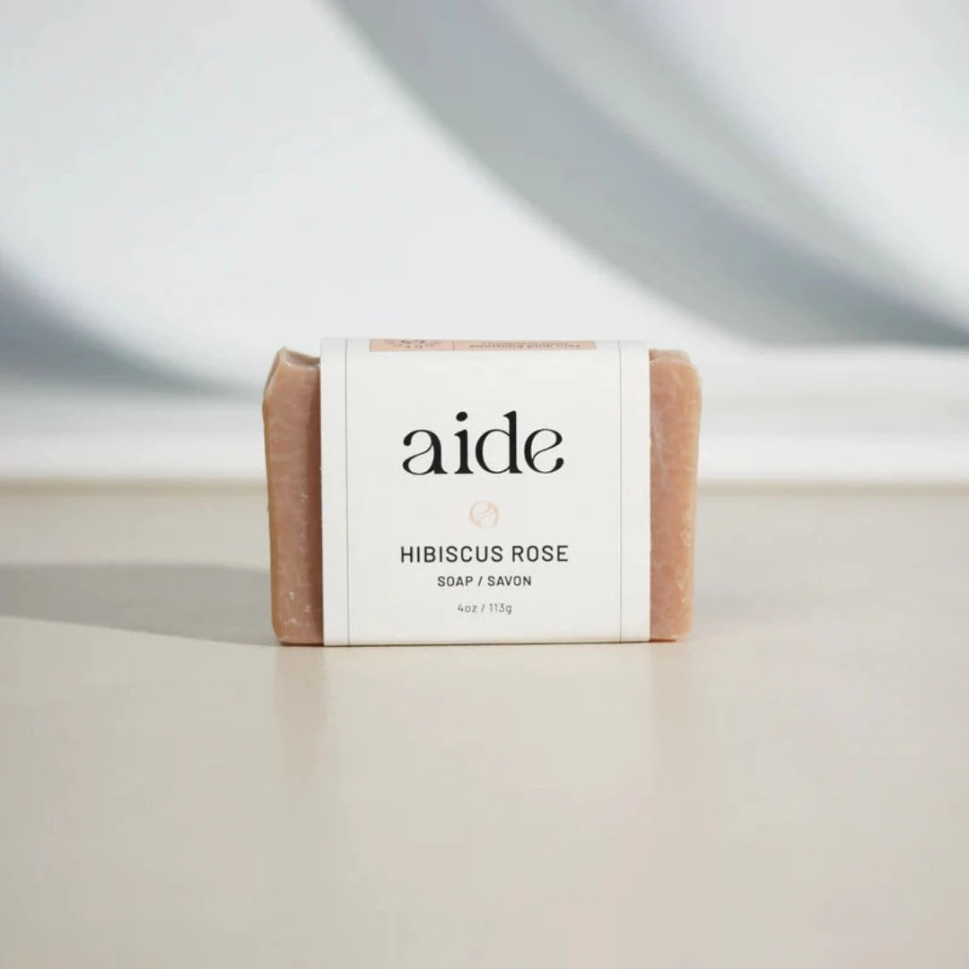 A bar of Aide Bodycare Hibiscus Rose Clay Soap, featuring a light pink color with a subtle texture and a paper label with the 'aide' logo and 'Hibiscus Rose' text, displayed on a light-colored surface with a shadow overlay. The soap is natural and handmade, available in Ottawa, Ontario, Canada.