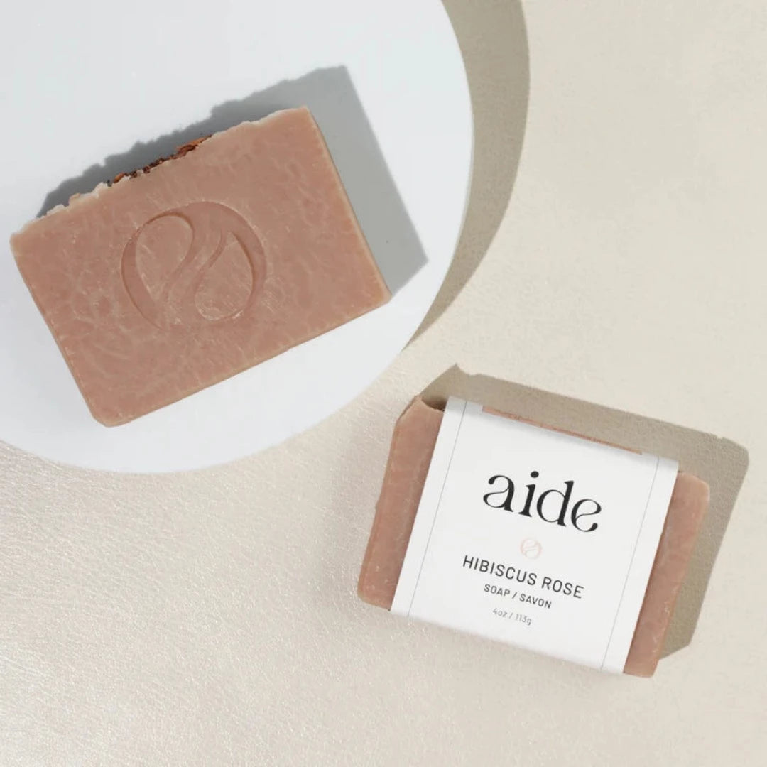 A bar of Aide Bodycare Hibiscus Rose Clay Soap, featuring a light pink color with a subtle texture and a paper label with the 'aide' logo and 'Hibiscus Rose' text, displayed on a light-colored surface with a shadow overlay. The soap is natural and handmade, available in Ottawa, Ontario, Canada.