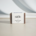 A bar of Aide Bodycare Lavender Oatmeal Soap, featuring a light purple-gray color with visible oatmeal texture and a paper label with the 'aide' logo and 'Lavender Oatmeal' text, displayed on a light-colored surface. The soap is natural and handmade, available in Ottawa, Ontario, Canada.