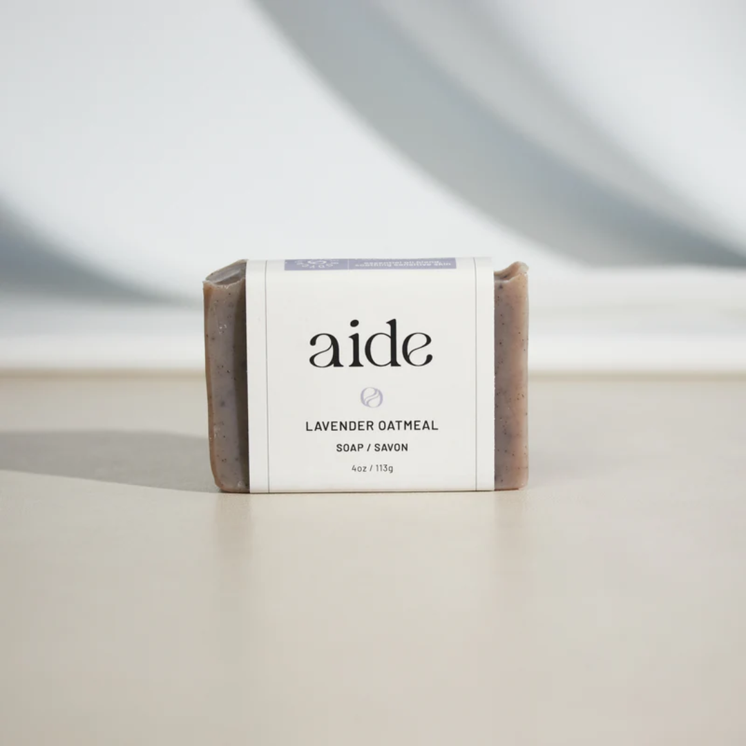 A bar of Aide Bodycare Lavender Oatmeal Soap, featuring a light purple-gray color with visible oatmeal texture and a paper label with the 'aide' logo and 'Lavender Oatmeal' text, displayed on a light-colored surface. The soap is natural and handmade, available in Ottawa, Ontario, Canada.