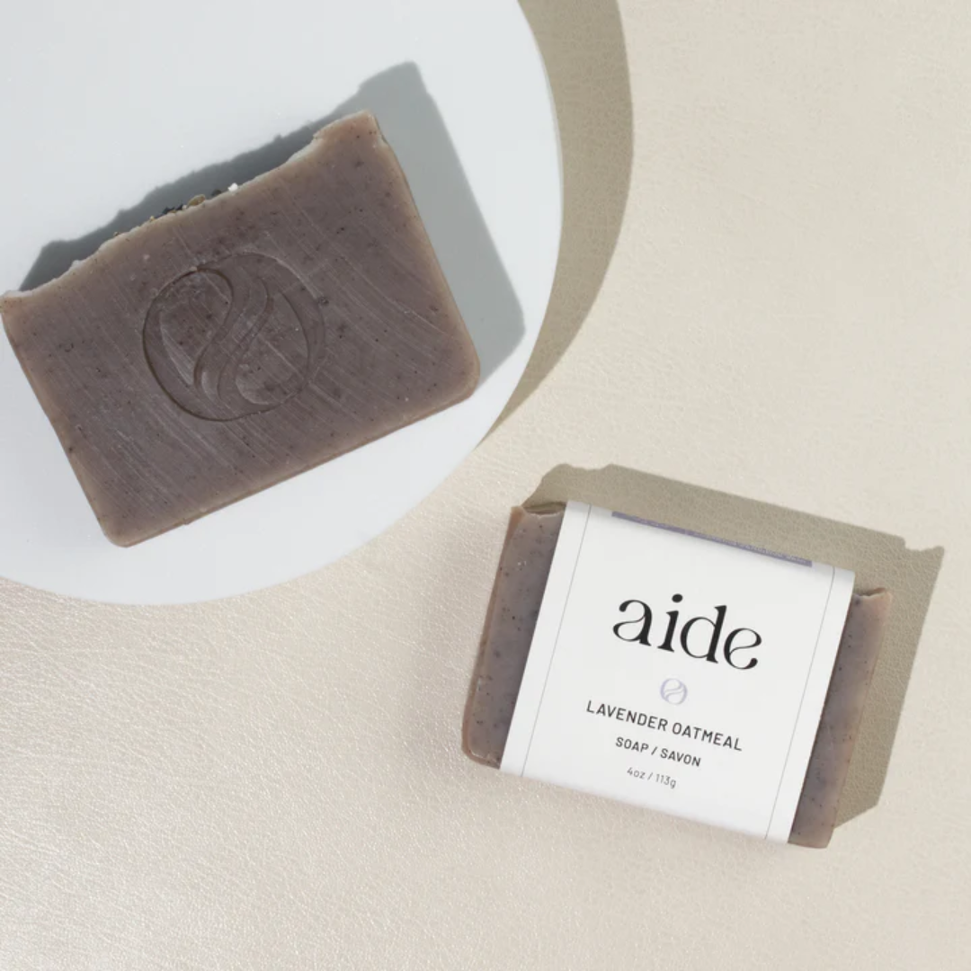 A bar of Aide Bodycare Lavender Oatmeal Soap, featuring a light purple-gray color with visible oatmeal texture and a paper label with the 'aide' logo and 'Lavender Oatmeal' text, displayed on a light-colored surface. The soap is natural and handmade, available in Ottawa, Ontario, Canada.