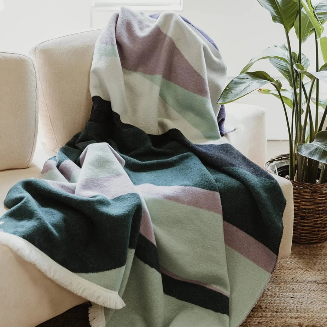 The Aurora Reversible Eco-Friendly Blanket by Mini Tipi, featuring flowing waves of green, purple, black, and white, draped on a sofa. This reversible and eco-conscious blanket is available in Ottawa, Ontario, Canada.