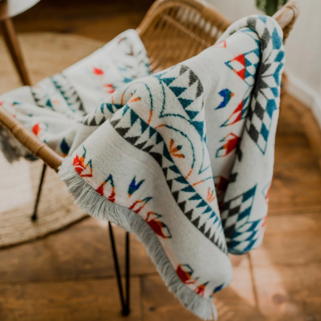 The Nibi Reversible Blanket by Mini Tipi, featuring bold Indigenous-inspired geometric patterns in teal, red, brown, and white, hanging from a chair. This reversible and stylish blanket is available in Ottawa, Ontario, Canada.
