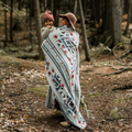 The Nibi Reversible Blanket by Mini Tipi, featuring bold Indigenous-inspired geometric patterns in teal, red, brown, and white, draped on a shoulders of a person holding a baby. This reversible and stylish blanket is available in Ottawa, Ontario, Canada.