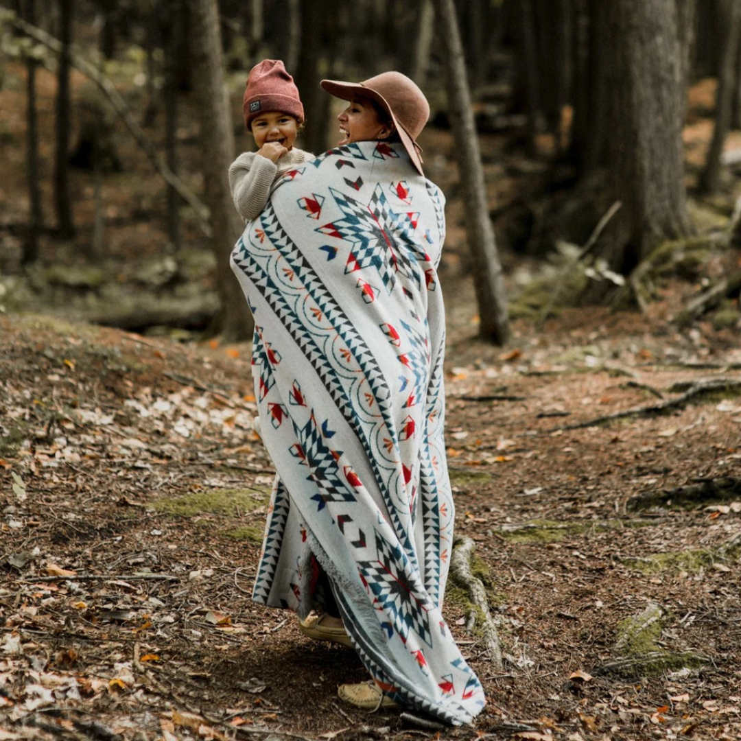 The Nibi Reversible Blanket by Mini Tipi, featuring bold Indigenous-inspired geometric patterns in teal, red, brown, and white, draped on a shoulders of a person holding a baby. This reversible and stylish blanket is available in Ottawa, Ontario, Canada.