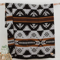 The Thunderbird Reversible Eco-Friendly Blanket by Mini Tipi, featuring bold Indigenous-inspired Thunderbird and geometric patterns in black, grey, and brown, hanging from a natural branch. This sustainable and culturally significant blanket is available in Ottawa, Ontario, Canada.