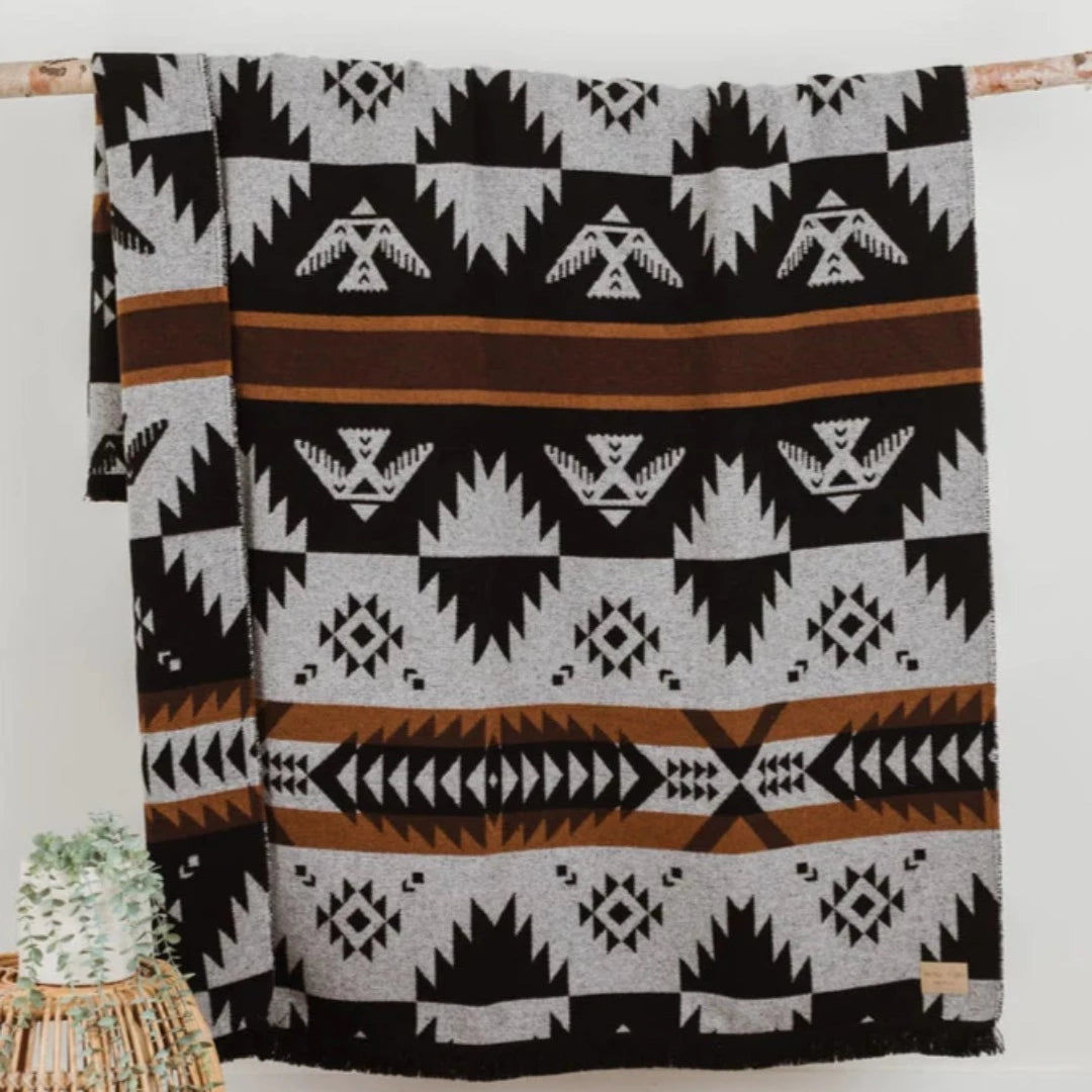 The Thunderbird Reversible Eco-Friendly Blanket by Mini Tipi, featuring bold Indigenous-inspired Thunderbird and geometric patterns in black, grey, and brown, hanging from a natural branch. This sustainable and culturally significant blanket is available in Ottawa, Ontario, Canada.