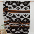 The Thunderbird Reversible Eco-Friendly Blanket by Mini Tipi, featuring bold Indigenous-inspired Thunderbird and geometric patterns in black, grey, and brown, hanging from a natural branch. This sustainable and culturally significant blanket is available in Ottawa, Ontario, Canada.