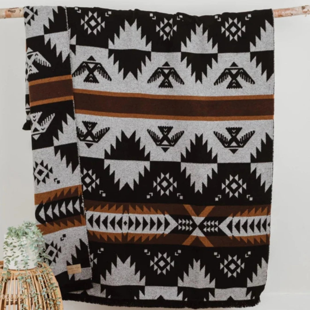 The Thunderbird Reversible Eco-Friendly Blanket by Mini Tipi, featuring bold Indigenous-inspired Thunderbird and geometric patterns in black, grey, and brown, hanging from a natural branch. This sustainable and culturally significant blanket is available in Ottawa, Ontario, Canada.