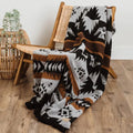 The Thunderbird Reversible Eco-Friendly Blanket by Mini Tipi, featuring bold Indigenous-inspired Thunderbird and geometric patterns in black, grey, and brown, handing from a wood chair. This sustainable and culturally significant blanket is available in Ottawa, Ontario, Canada.