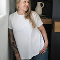 Woman wearing a white t-shirt and blue jeans standing indoors with a shelf and decor items in the background.