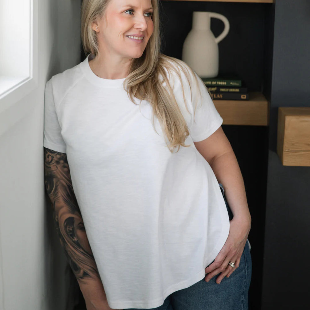 Woman wearing a white t-shirt and blue jeans standing indoors with a shelf and decor items in the background.
