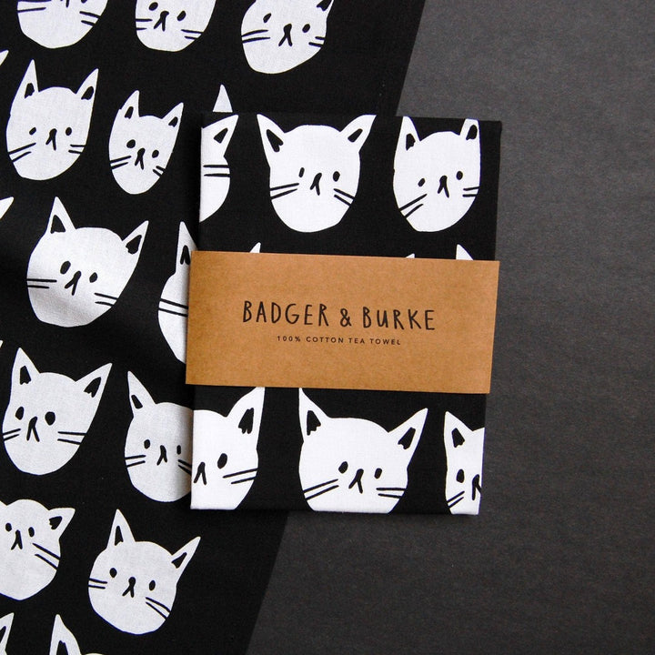 A black tea towel with a pattern of white cat faces. The cat faces are arranged in a grid-like pattern and vary in size and shape.